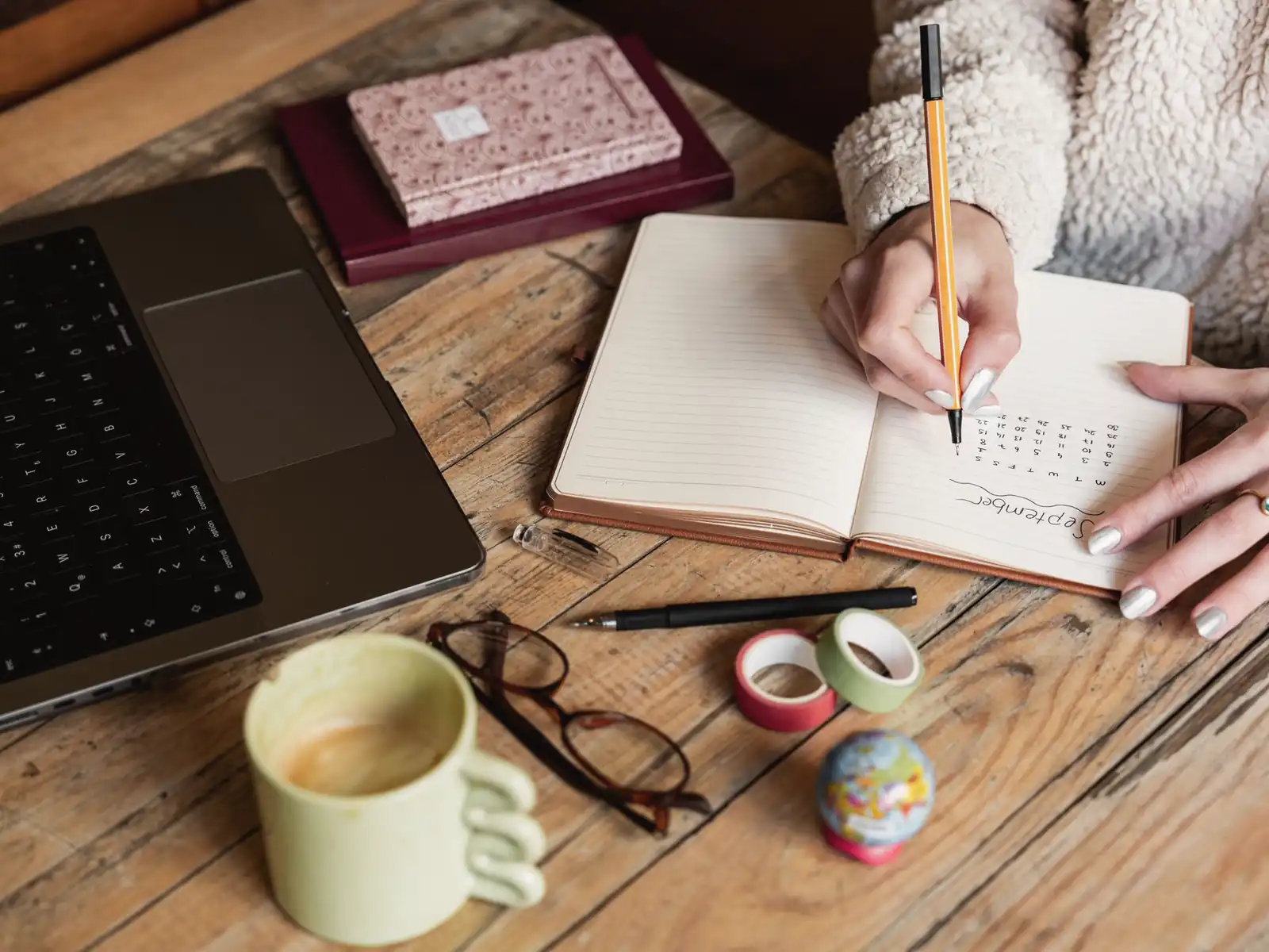 Hands writing in a paper planner beside a laptop on a wooden table
